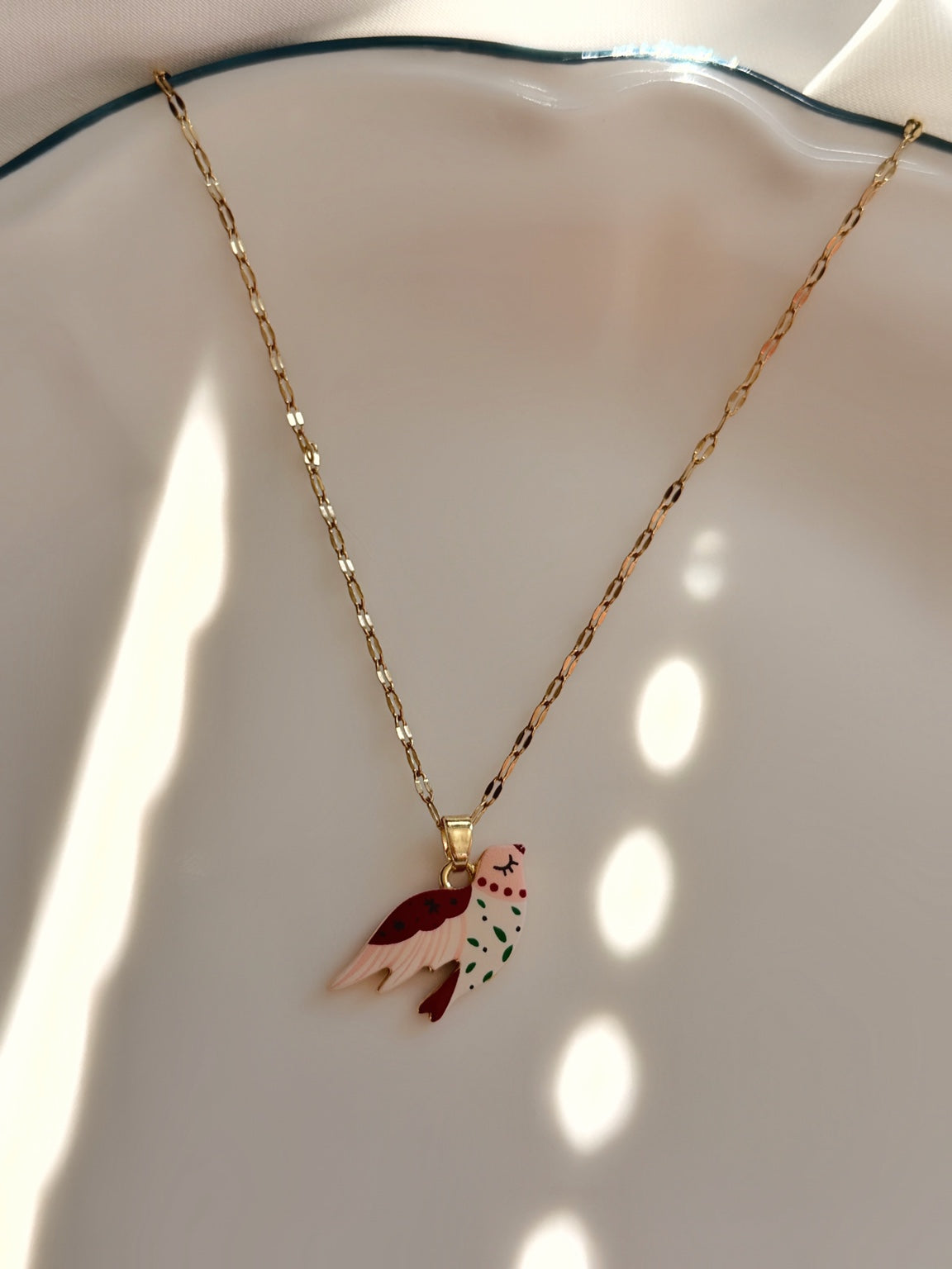Beyaz porselen tabakta gold tonlu zinciriyle sergilenen kırmızı beyaz mineli kuş figürlü kadın kolye.