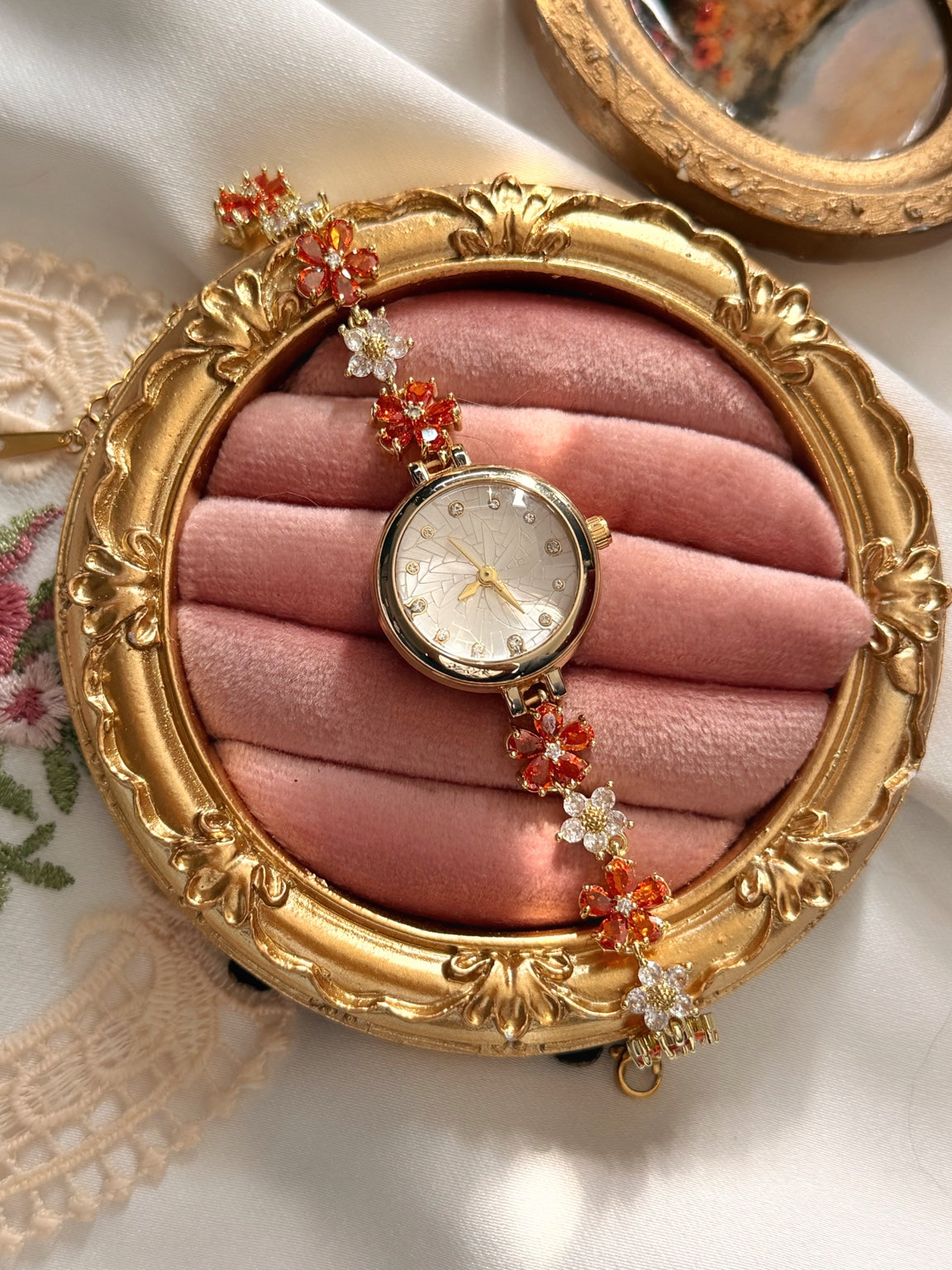 Pembe kadife takı standında kırmızı çiçekli kadın saatin desenli açık renk kadranı, gold kasası ve taşlı kordon detayları.