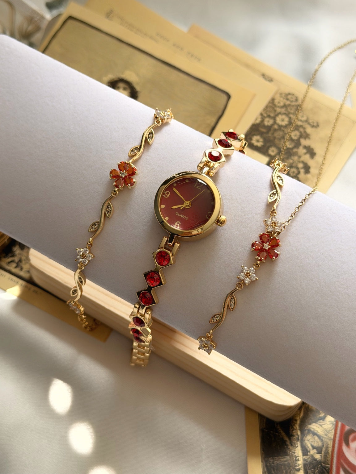 Krem stand üzerinde kırmızı taşlı gold saat, kırmızı çiçekli çelik kolye ve bileklikten oluşan üçlü kadın takı seti