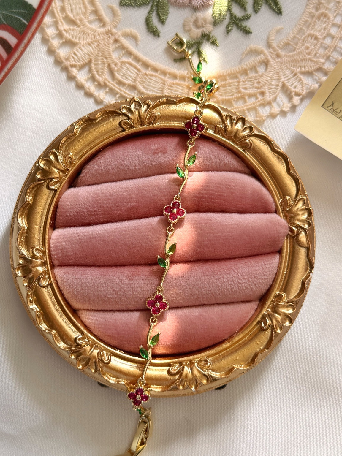 Pembe kadife üzerinde kırmızı yonca motifleri, yeşil yaprak taşları ve gold bağlantıları görülen kadın bileklik.