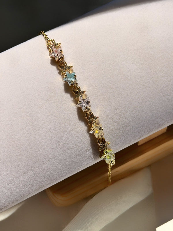 Beyaz kadife bileklik standında pastel yıldız taşları ve ince gold zincir, gün ışığında çapraz görünüm.