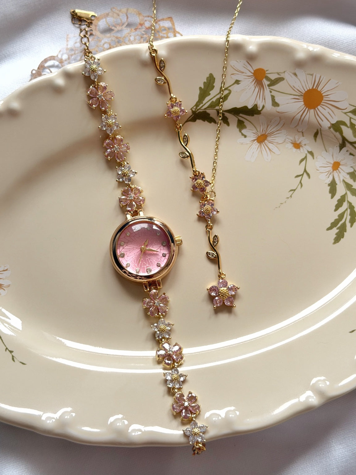 Pembe kadranlı çiçekli gold kadın saat ve Y kolye takı setinin çiçek desenli tabak üzerinde görünümü