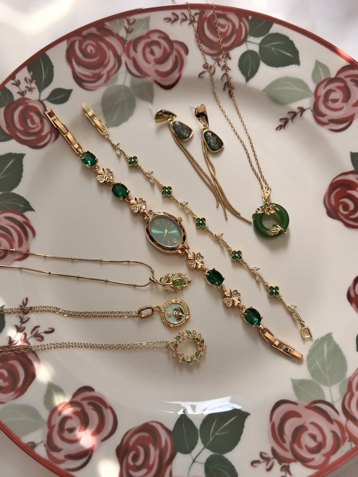 Çiçek desenli tabakta yeşil ve gold tonlu 7'li takı setinin farklı açıdan görünümü; dört kolye, oval saat, yonca bileklik ve bir çift küpe.