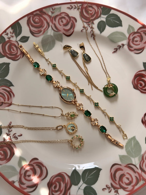 Çiçek desenli tabakta yeşil ve gold tonlu 7'li takı setinin farklı açıdan görünümü; dört kolye, oval saat, yonca bileklik ve bir çift küpe.